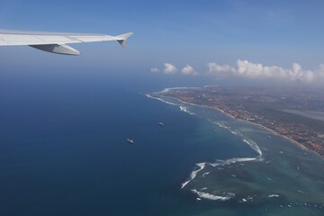 Fototapeta premium Aerial view from airplane window of Bali's coastline, featuring blue ocean, sandy beaches, and buildings. Airplane wing visible, indicating photo taken during flight.