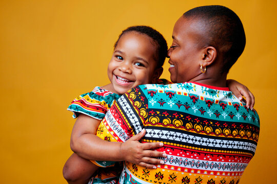 Black woman holding smiling Black child, both wearing patterned clothing, child looking at camera while woman looking at child, close interaction against plain yellow background - Powered by Adobe