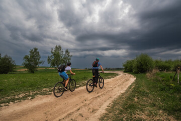 Two cyclists riding on a sandy rural road under heavy storm clouds in the countryside of Małopolska, Poland. Dynamic weather and open fields with scattered trees.
