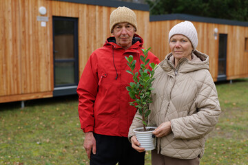 Two individuals wearing warm clothing hold a potted plant outside modern wooden cabins. The scene reflects a peaceful atmosphere in a natural setting
