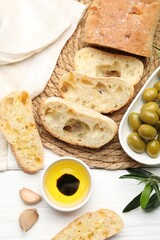 Fresh cut ciabatta, oil, olives, garlic and leaves on white wooden table, flat lay