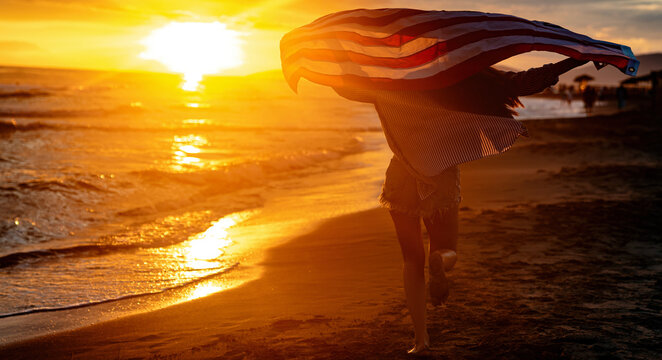 Young woman holding national American flag walking ocean beach. America Independence Day concept