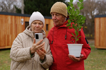 Two seniors are enjoying a moment together outside, focused on a phone screen while one holds a potted plant, surrounded by wooden sheds in a garden