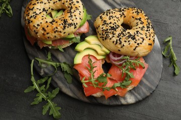Delicious bagels with salmon, avocado, onion and arugula on black table, flat lay