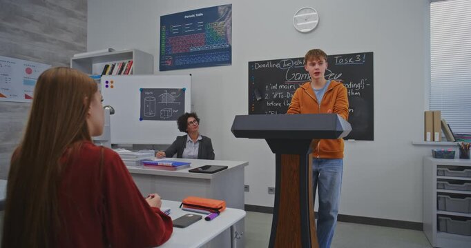 Focused Boy in Bright Orange Hoodie Delivers Class Presentation on Creative School Project. Teacher and Students Listen Attentively. Concept Creativity, Communication, and Collaboration in Education.