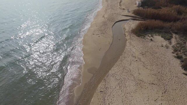 Aerial view of sandy beach with small river flowing into the sea. Coastal landscape with gentle waves, shoreline, and natural water patterns on a cloudy day.