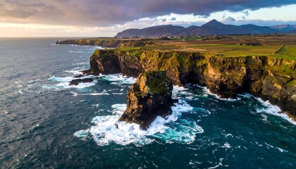 Rocky coastline with cliffs and mountain backdrop under cloudy sky, ocean waves crashing