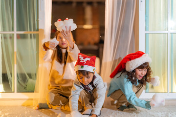 Three children wearing Christmas accessories sit at the doorway holding artificial snow, enjoying the holiday spirit with joy and curiosity in a cozy indoor winter-themed setting.
