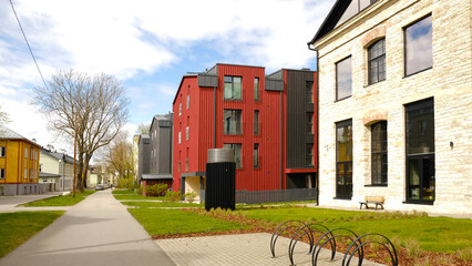 Modern red and grey residential buildings and a renovated brick house. A tranquil urban street with green lawns and bike rack. Contemporary city life scene. City living and real estate.