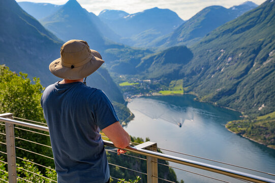 Tourist wearing a sun hat enjoys panoramic view of Geiranger Fjord from a scenic lookout in Norway.