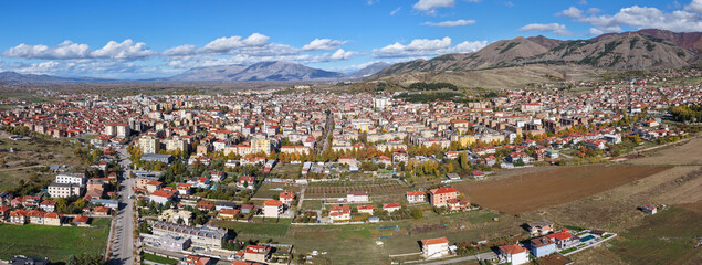 Panoramic autumn aerial of Korçë, showcasing the city’s dense layout, open fields, and dramatic mountain backdrop under clear blue skies.