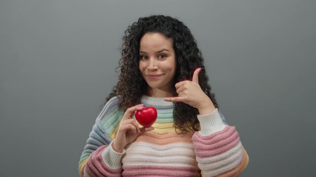 Hispanic woman holding red heart and gesturing call me against grey background wearing colorful sweater, showing joy and communication.