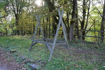 An Old Weathered Wooden Frame Stands In A Sunlit Grassy Clearing Among Autumn Trees And A Fence At Black Rocks, Cromford, Derbyshire.