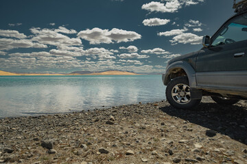 An off-road vehicle with mud tires parked on the rocky shore of a pristine blue lake in the Mongolian desert. Sand dunes and cloudy blue sky in the distance. Tire-focused composition.