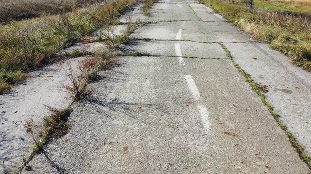 Abandoned asphalt road. Grass breaks through and grows from cracks in the asphalt. Damaged cracked road path with weeds growing. Cracked asphalt road with weeds breaking through.