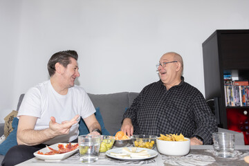 Mature male couple laughing and gesturing while eating together at home. Close domestic moment, horizontal with copy space. Concept of friendship and daily life