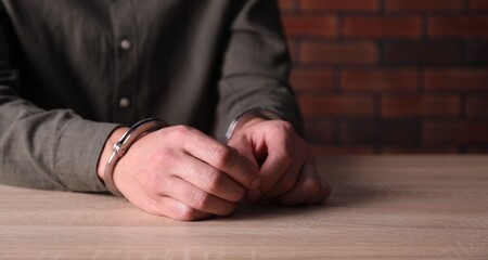 Man in handcuffs at wooden desk indoors, closeup. Space for text