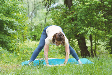 Woman practicing yoga in a wide-legged forward fold on a mat outdoors amidst lush green nature. Embracing wellness and mindful movement in a serene natural setting.
