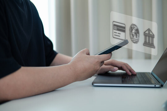 Fintech concept of a person using a smartphone and laptop for digital banking. Hand holds phone with virtual icons for online payment, global e-commerce, and secure financial transactions