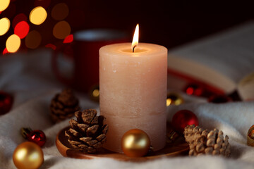 Burning candle, cones and baubles on light blanket against black background with blurred lights, closeup. Bokeh effect