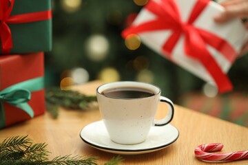 Woman holding gift box and wooden table with aromatic coffee, candy cane and fir tree branches against blurred lights, selective focus. Christmas greeting card