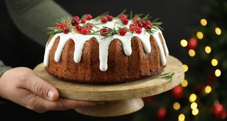 Woman holding delicious Christmas cake with icing, cranberries and rosemary against black background with blurred lights, closeup. Bokeh effect