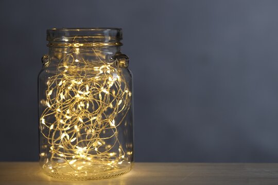 Christmas lantern. Glowing fairy lights in glass jar on table against grey background, space for text - Powered by Adobe