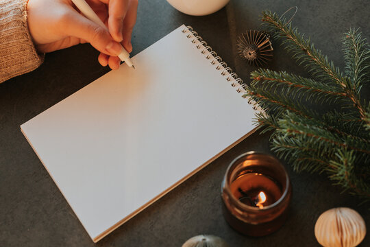 Female hand writing in notebook. Fir tree, burning candle, cup of coffee and decoration on table. Top view, flat lay. New year's Resolution and Goals