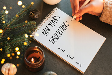 Woman's hand writing New Year's Resolution's in notebook. Fir tree, candle, cup of coffee and decoration on table. Top view, flat lay. Festive Mood Atmosphere