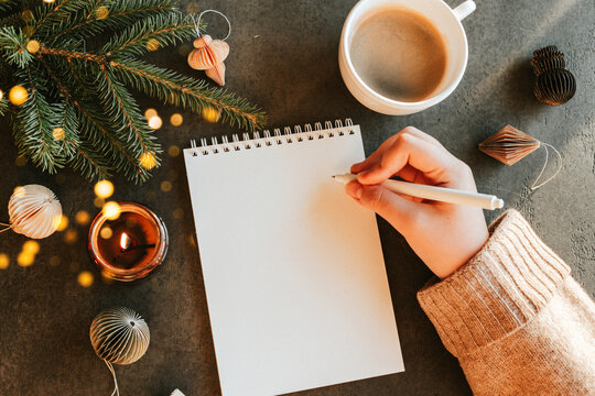 Female hand writing in notebook. Fir tree, burning candle, cup of coffee and decoration on table. Top view, flat lay. New year's Resolution and Goals - Powered by Adobe