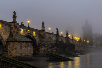 Fototapeta premium Charles Bridge over the Vltava River in Prague, Czech Republic, Shrouded in Morning Fog at Sunrise with Glowing Street Lamps, Empty of People, Creating a Calm and Atmospheric Cityscape