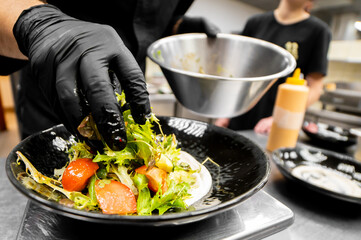 A chef in black gloves precisely plates a fresh green salad with tomatoes and other ingredients in a professional kitchen setting.