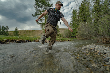 A traveler runs through a mountain stream on a green meadow in the Altai steppe. Demonstrating waterproof footwear with pine trees and a hill in the background.

