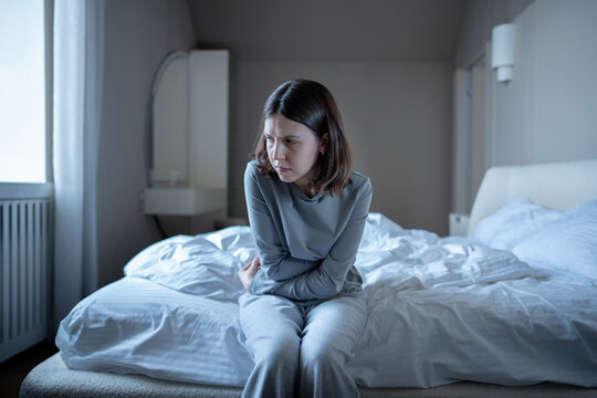 Stressed unwell woman sits hunched on unmade bed, arms crossed over stomach struggles with painful cramps. Anxious frustrated young female alone in bedroom deep in depressed thought, mental exhaustion - Powered by Adobe