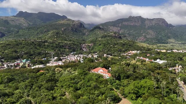 Mesmerising aerial footage of a lush South Indian town nestled at the foothills of the Western Ghats, surrounded by dense greenery and towering cloud-clad moving mountains.