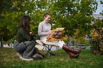 Two women talking while painting outdoors in park.