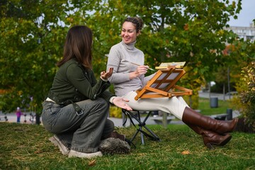 Two women talking while painting outdoors in park.