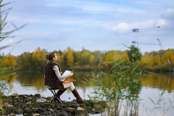 Woman painting outdoors by autumn lake on rocky shore.