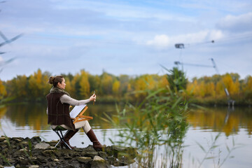 Woman painting outdoors by autumn lake on rocky shore.