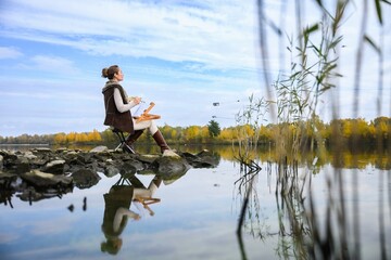 Woman painting outdoors by autumn lake on rocky shore.