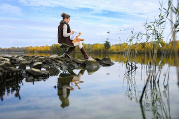 Woman painting outdoors by autumn lake on rocky shore.