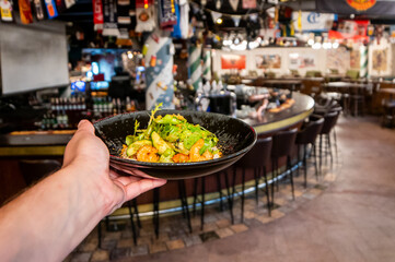 A person's hand holding a black bowl filled with a fresh, vibrant shrimp and cucumber salad with lettuce greens, served in a dimly lit, casual bar or restaurant interior