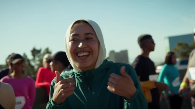 A young woman wearing a hijab smiles and gives thumbs up at a running event. She stands among other participants, radiating joy and enthusiasm for fitness.