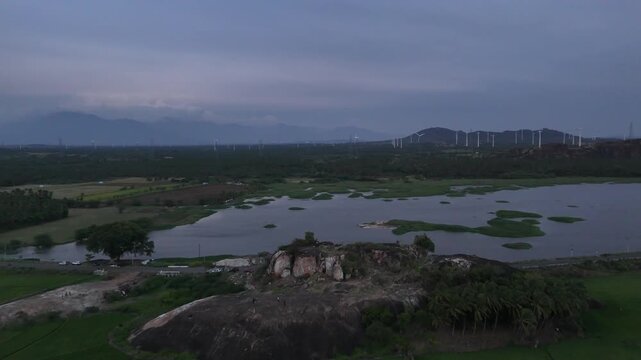 Aerial view of a rural landscape at dusk. A lake sits in the foreground, with a wind farm visible on the hills in the background. A distant mountain range frames the scene under a moody, overcast sky.