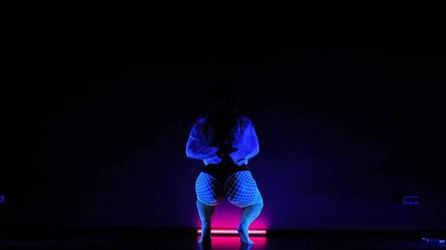 A young woman dances twerk in fishnet tights under neon lights. 