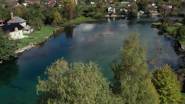 Aerial footage of the Una River near Bihac, Bosnia and Herzegovina. Calm clear water flows beside riverside houses, trees, and gardens, showing peaceful nature and local village scenery.