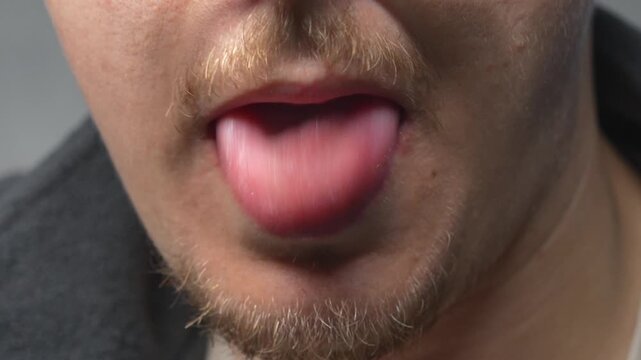 A man sticks his tongue out at the camera against a gray background.
Wet tongue in a man's mouth.
Male facial expression and obscene gesture.