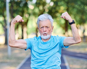 Portrait of a happy positive old senior man showing biceps and power standing outdoors in park or nature, healthy lifestyle, active senior, vitality and humor concepts