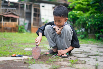 A young boy sits in the yard while digging soil using a small gardening shovel, playing with toy cars on the ground. The scene captures a candid outdoor moment of childhood play and creativity.