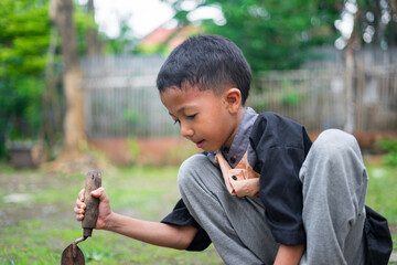 A young boy concentrates while holding a small gardening shovel during outdoor playtime. He sits on the grass in a natural backyard environment, enjoying simple hands-on activities.